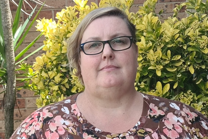 close up image of a woman with short blonde hair and black glasses, looking straight ahead