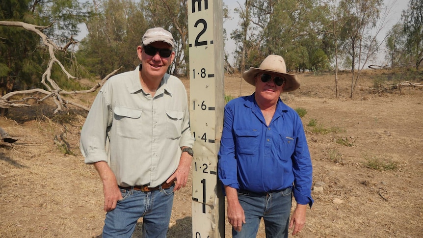 Phil and Rick Laird standing either side of a water level marker in a dry river bed.