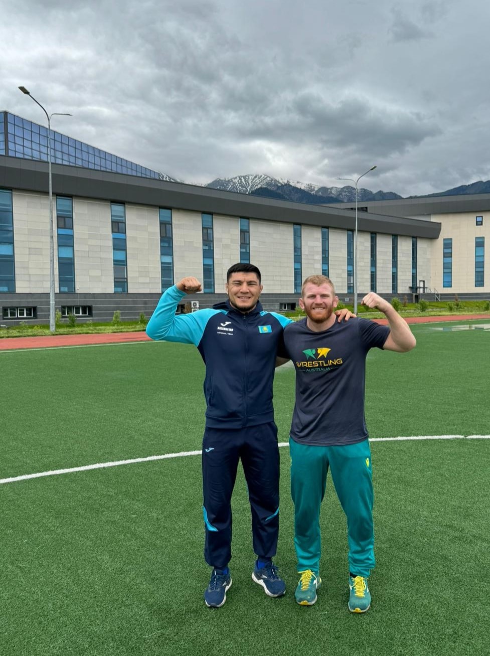 Two men stand on a soccer pitch, arm in arm, flexing their muscles. Both smile. Building and mountains behind them.