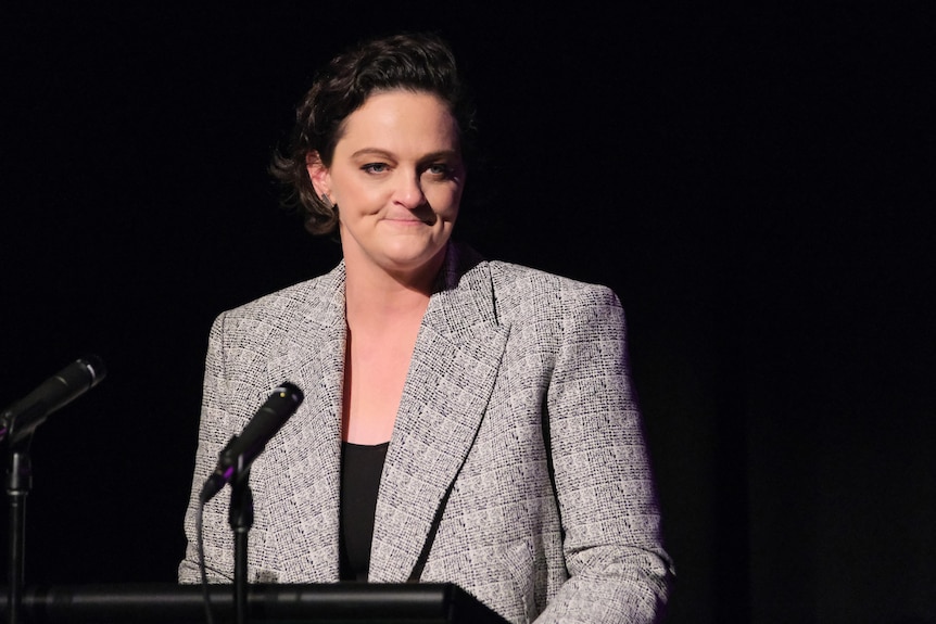 A woman wearing a grey suit speaks at a podium