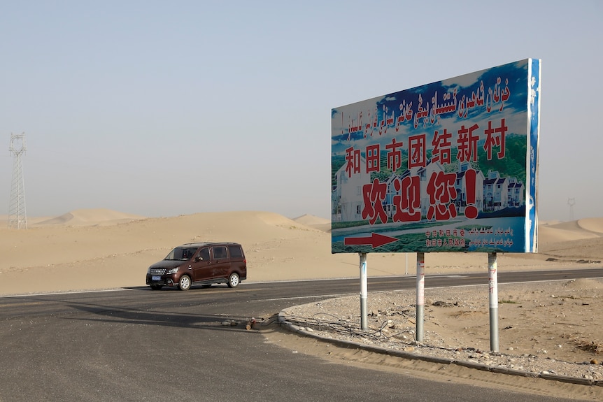 A car drives through a desert where a signboard which reads "Welcome to the Hotan Unity New Village".