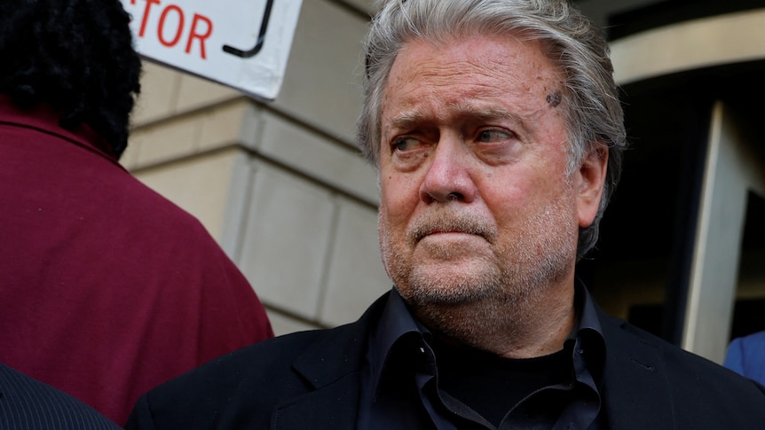 Steve Bannon is pictured wearing a black suit, looking to the left with a disappointed look on his face.