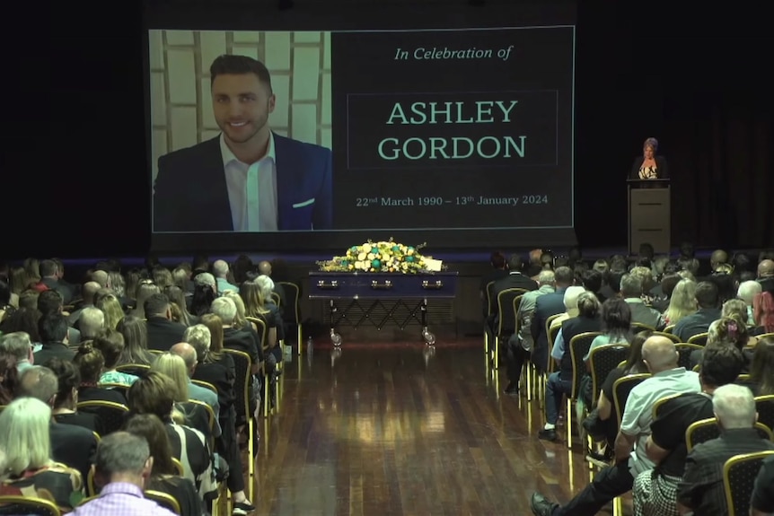 A funeral with mourners sitting in seats and picture of Ashley Gordon on screen.