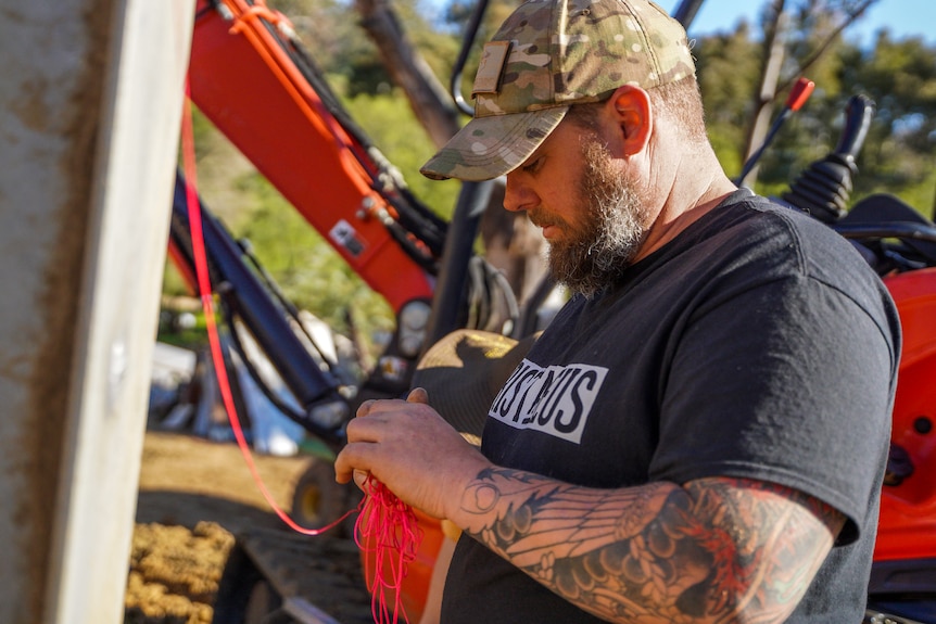 A bearded man wearing a cap and black shirt ties rope around a pole on a work site.