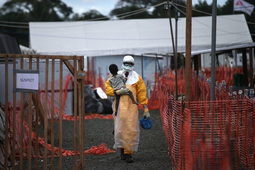 Wearing a bright yellow protective suit, a Ebola treatment centre staff member carries a small child from a white tent.