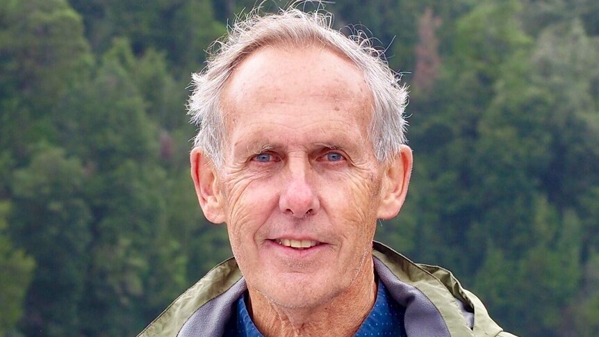 Bob Brown outside wearing a blue jumper and khaki jacket, standing in front of a river and trees.