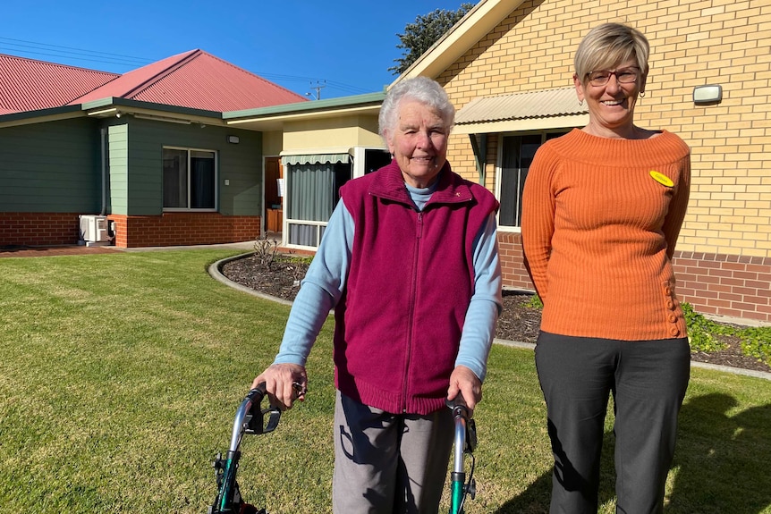 A middle-aged woman stands smiling alongside an older women bent over a walker in front of a brick residential unit.