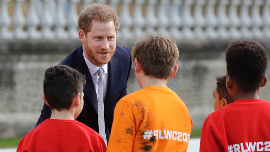 Prince Harry shakes the hand of schoolchildren who are wearing shirts emblazoned with "#RLWC2021".