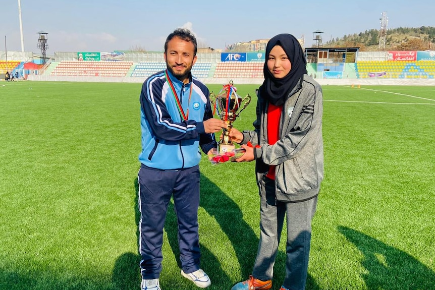 Adiba stands with her former Afghanistan soccer coach on a pitch holding a trophy.