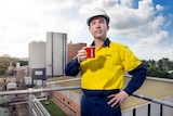 A mid shot of a man in high vis standing on a balcony holding a coffee cup with a factory in the background