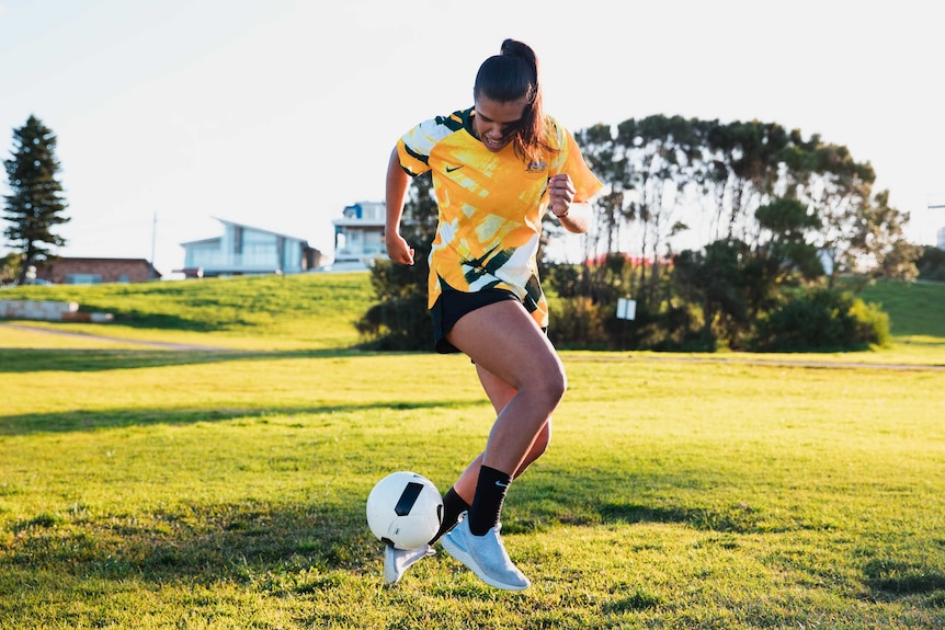 Jada Whyman kicks a white soccer ball outside in a park wearing a yellow soccer jersey, black shorts and back socks