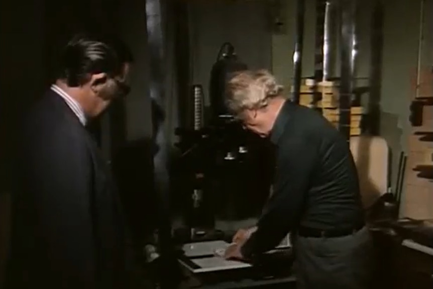 A 1970s still of two men looking at machinery in a basement