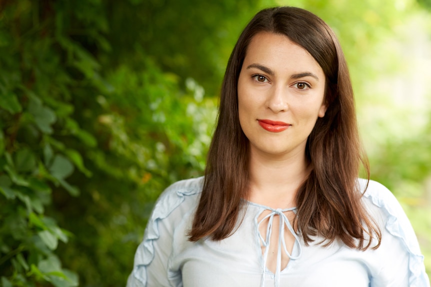 Playwright has long dark brown straight hair, and stands wearing light blue blouse in front of out-of-focus greenery outdoors.