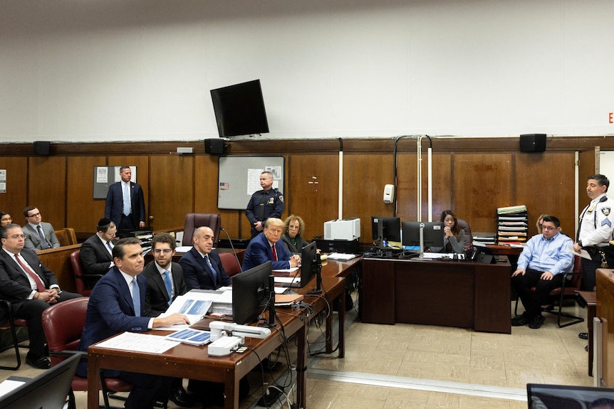 A wide shot of Donald Trump sitting at a bench in a court, with lawyers seated with him and guards throughout the room.