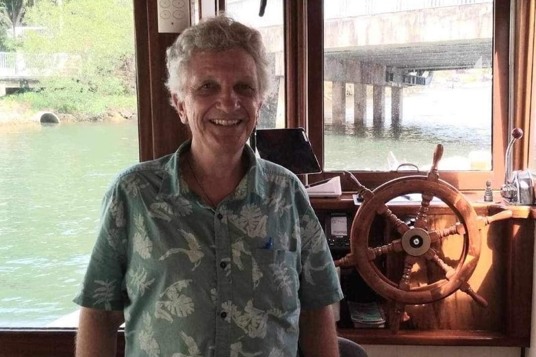 Man with grey hair smiles wearing patterned shirt in front of steering wheel of river boat