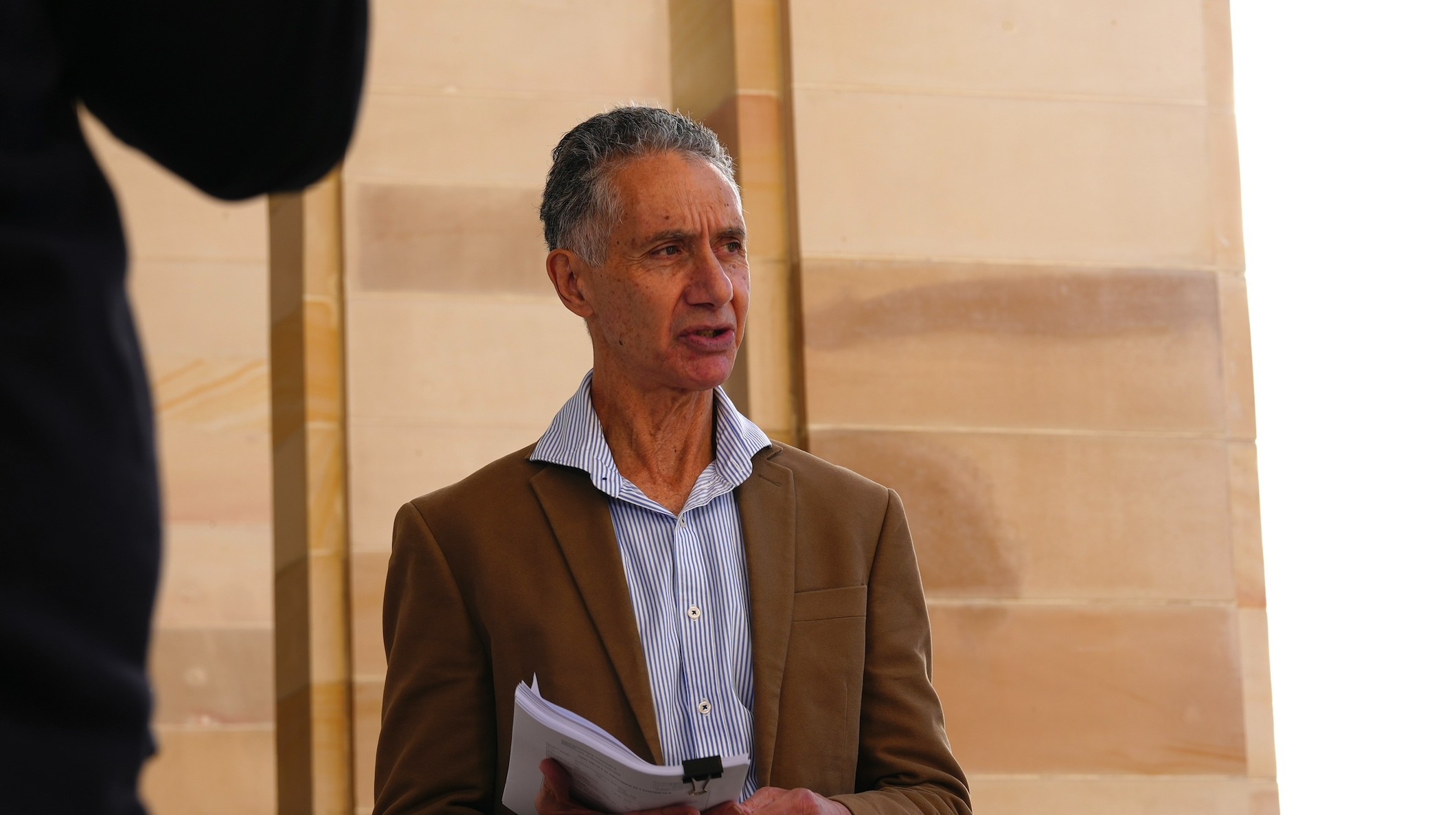 A mid-shot of a man wearing a brown suit jacket and light-coloured shirt 