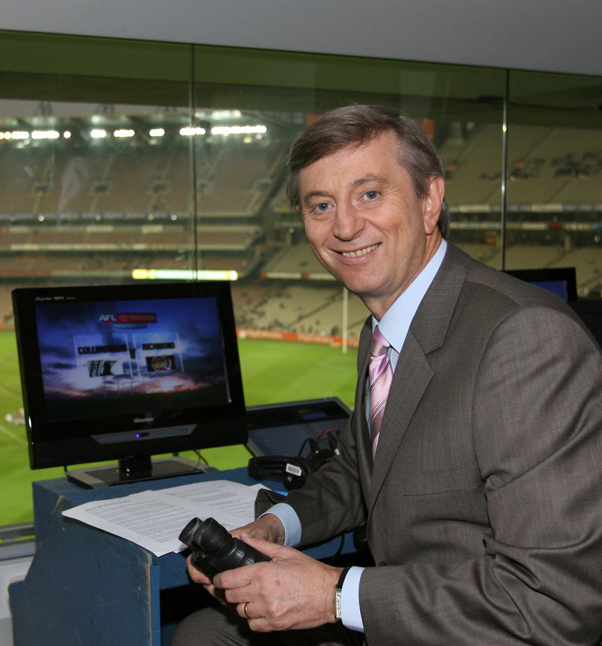 Um homem sorridente, de meia-idade, de terno, está sentado em uma cabine de comentários com vista para um campo esportivo em um estádio.