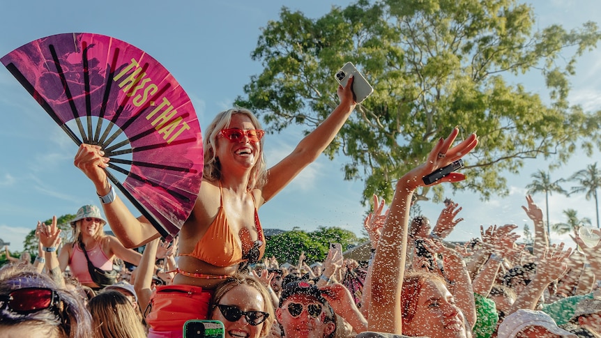 A festival-goer holding a pink fan atop shoulders in a packed crowd against a blue sky