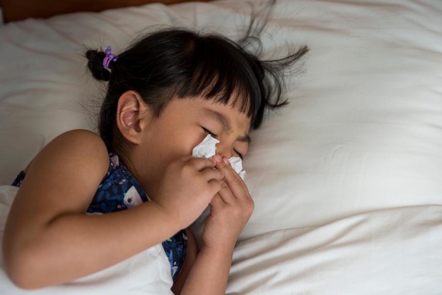 A young girl in a bed with white sheets blowing her nose. 
