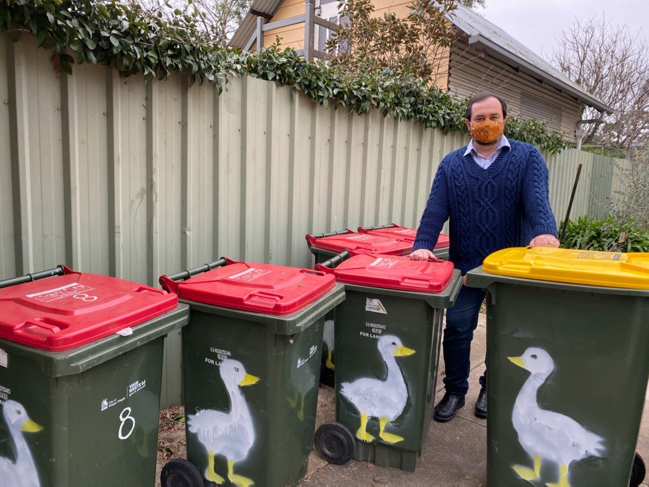 Man in knit navy jumper and orange face mask  standing behind kerbside bins featuring a painting of a goose on each one