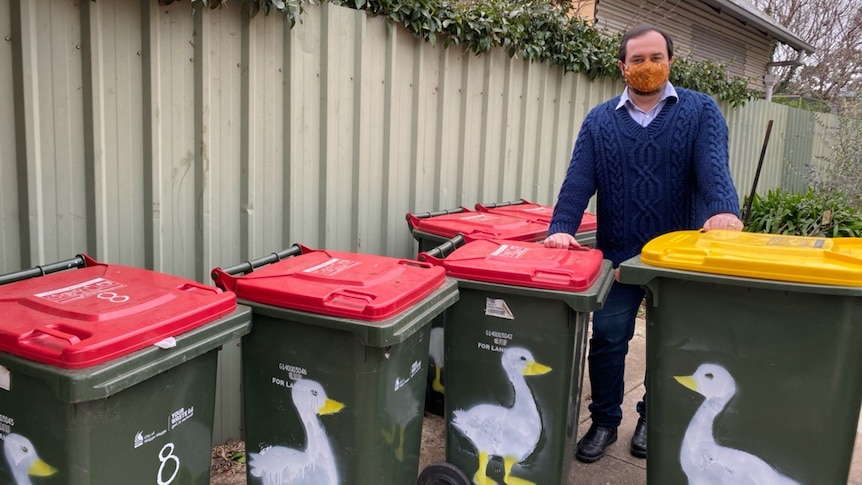 Man in knit navy jumper and orange face mask  standing behind kerbside bins featuring a painting of a goose on each one