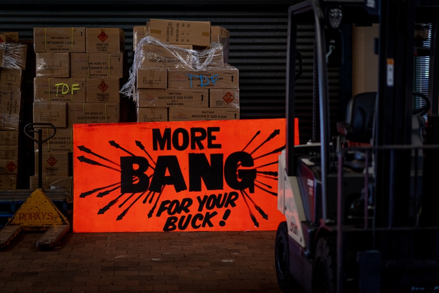Stacks of boxes inside a warehouse, behind a large bright orange sign reading "More Bang for your Buck".