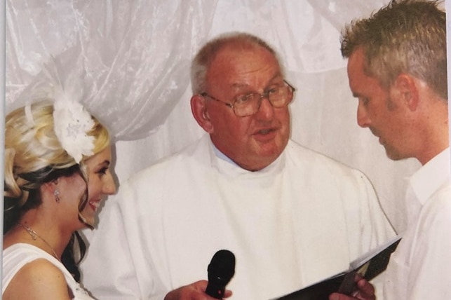 Des Williams speaks into a microphone behind the bride and groom.