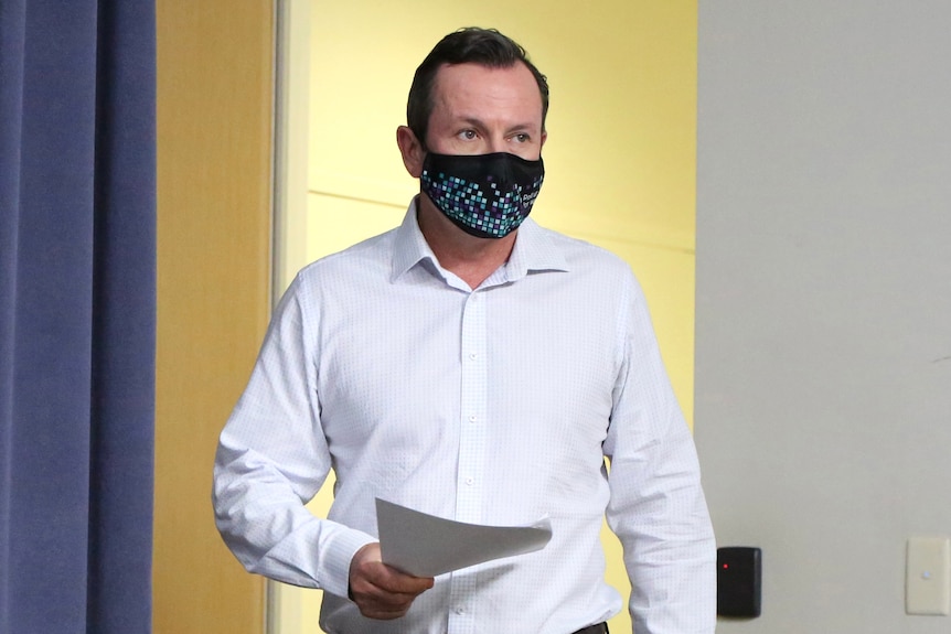 A mid-shot of WA Premier Mark McGowan walking wearing a white shirt, holding papers and wearing a face mask.