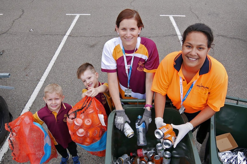 School students and two women stand behind wheelie bins filled with recyclable bottles
