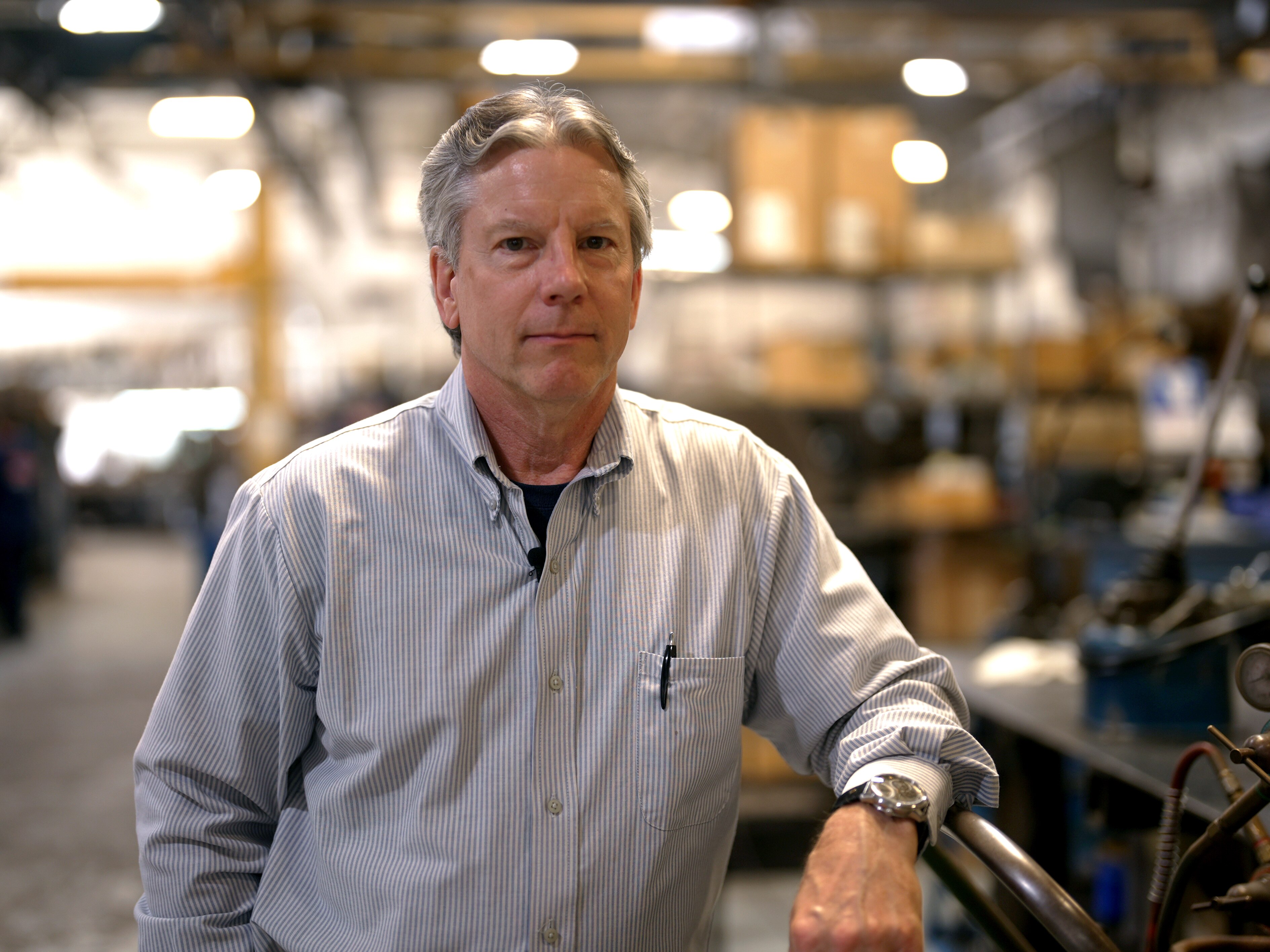 A man in a button up shirt leans against a bench. Behind him is a workshop. He has a neutral expresion.