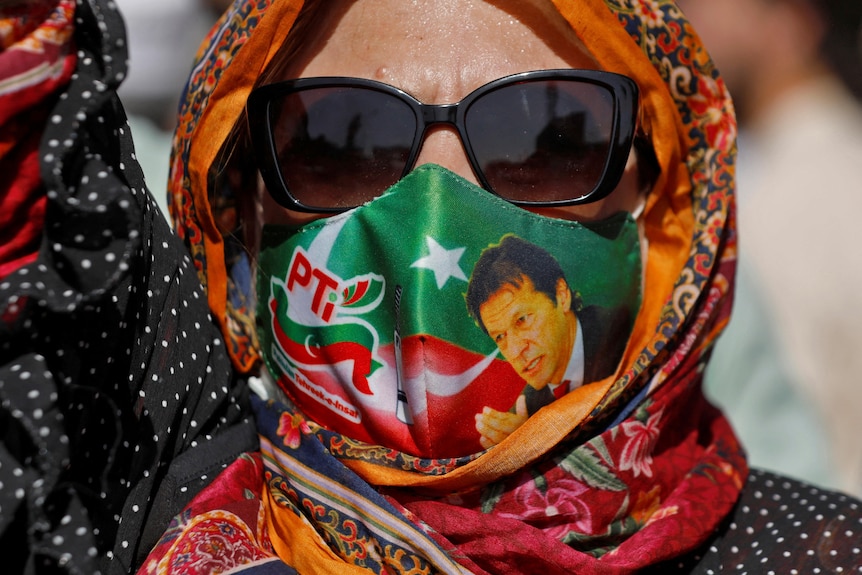 A woman in an Imran Khan face mask, sunglasses and a headscarf raises an arm