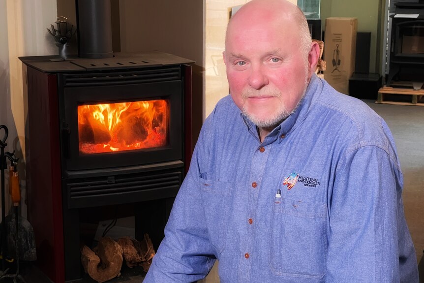 John sits in front of a wood heater, which is burning brightly. He has a silver beard and wears a shirt with a logo on it.