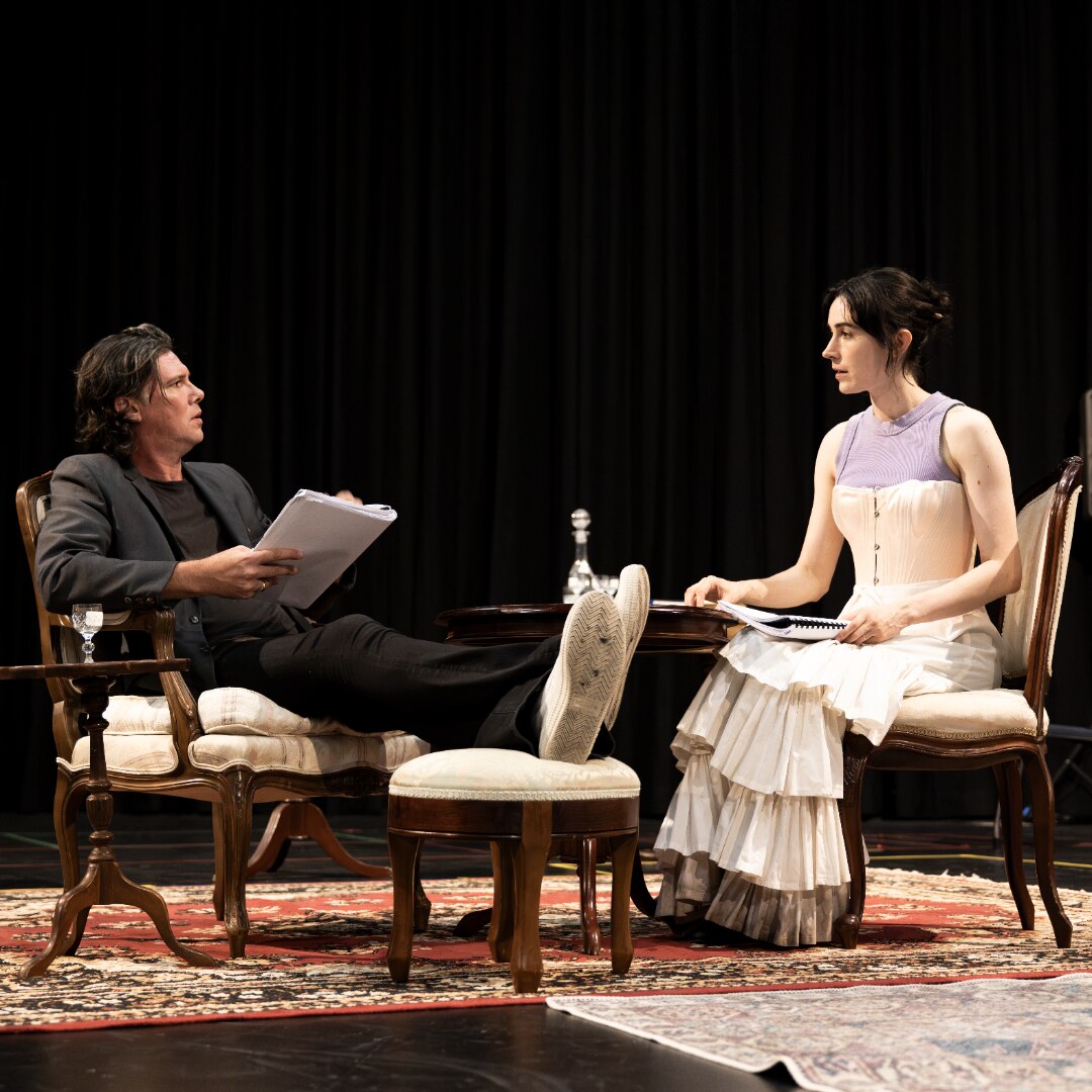 Toby Schmitz, a brunette white man, and Geraldine Hakewill, a brunette white woman, sit opposite each other rehearsing lines.