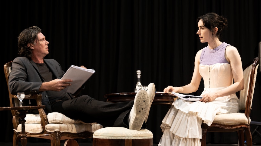 Toby Schmitz, a brunette white man, and Geraldine Hakewill, a brunette white woman, sit opposite each other rehearsing lines.