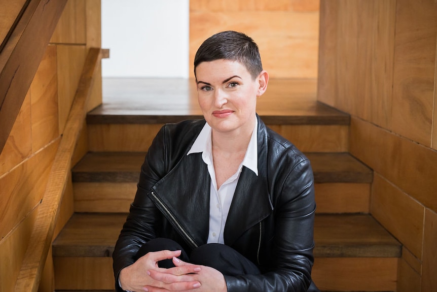 Martha with a shaved head and leather jacket, sitting down for a professional headshot.