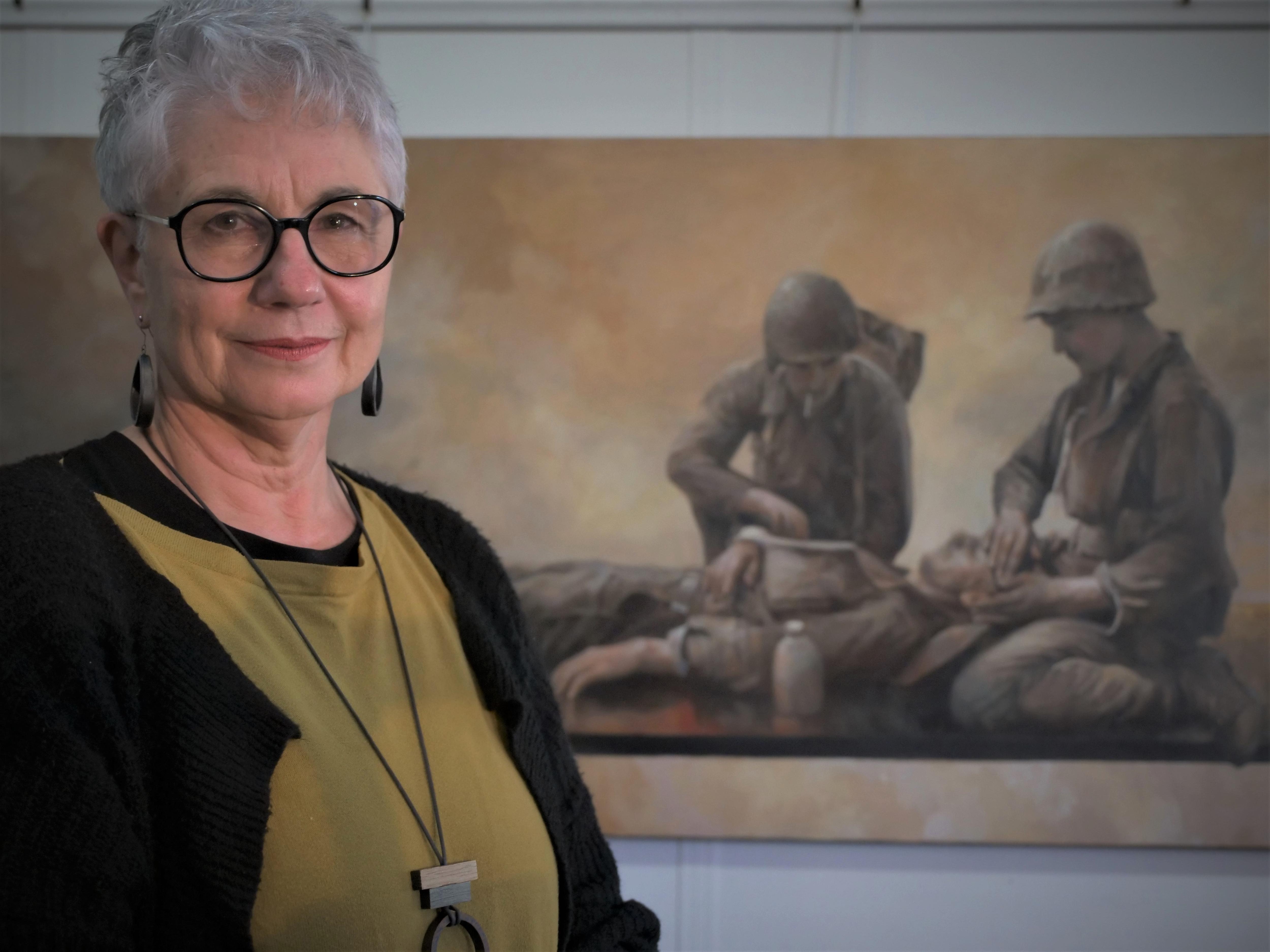 A woman with short hair and glasses standing in front of a large horizontal painting of soliders.