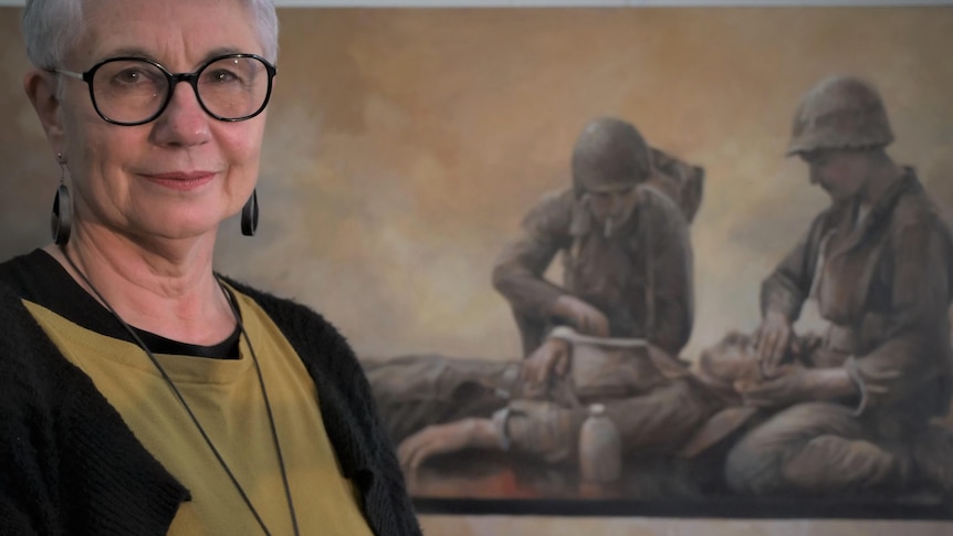 A woman with short hair and glasses standing in front of a large horizontal painting of soldiers