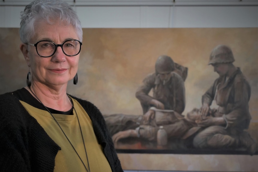 A woman with short hair and glasses standing in front of a large horizontal painting of soliders.