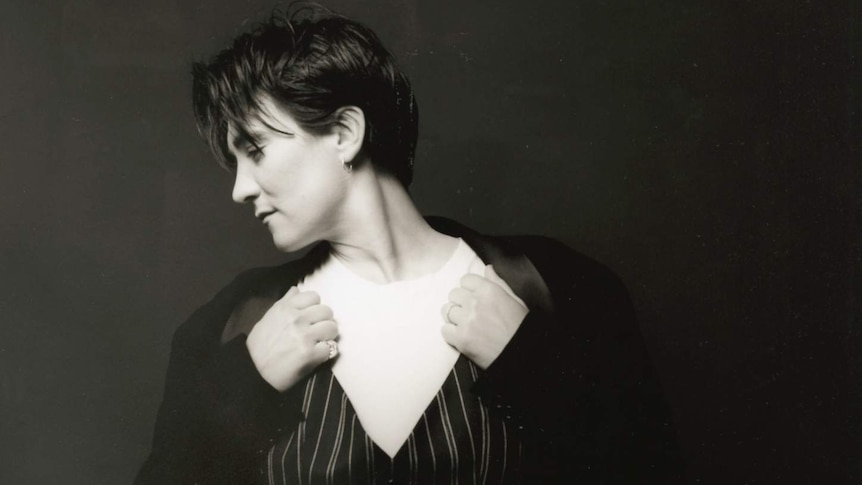 k.d. lang, in her thirties, looks to the left wearing a pinstriped suit over a white t-shirt against a dark background