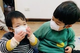 Two children, both wearing protective masks over their mouths, play with each other on the floor