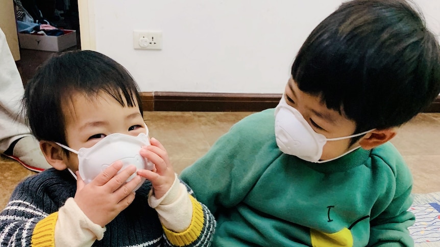 Two children, both wearing protective masks over their mouths, play with each other on the floor