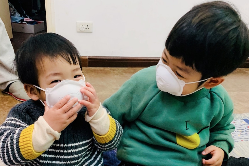 Two children, both wearing protective masks over their mouths, play with each other on the floor