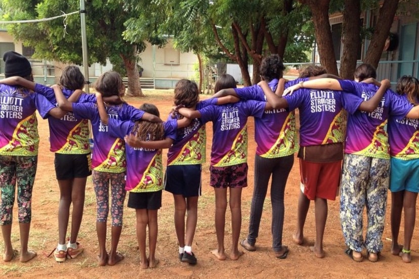 A group of young Indigenous girls all standing with their backs to the camera with their arms around each other with one adult.