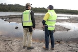 Two people wearing fluoro yellow vets stand in front of a stream of water and muddy land.