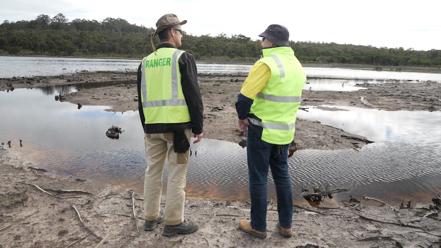 Two people wearing fluoro yellow vets stand in front of a stream of water and muddy land.