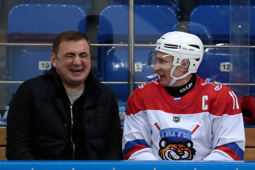 Two men, one who is Vladimir Putin wearing a helmet and ice hockey outfit, laugh together while sitting side by side
