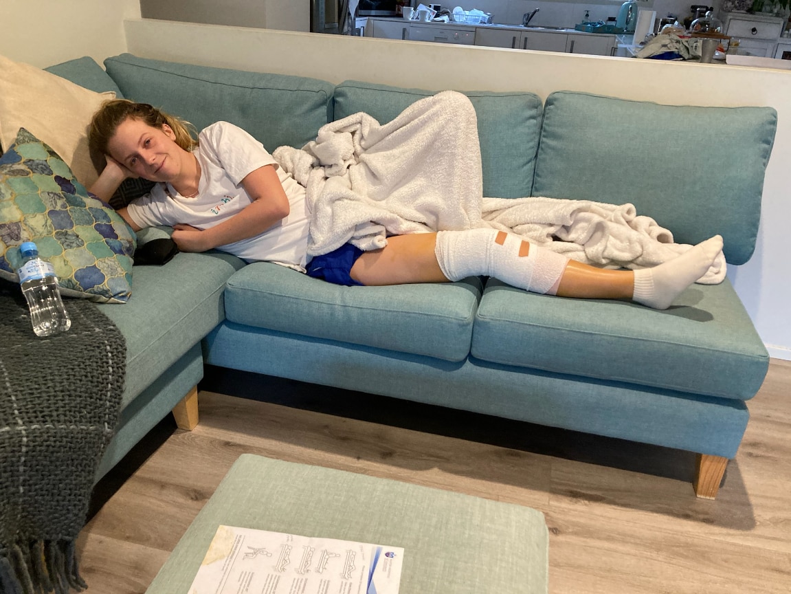 A blonde woman in her 20s, Annaliese Shaw, lies on a couch with a leg brace in some active wear and a blanket.