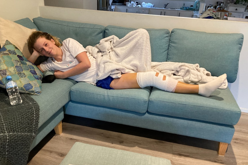A blonde woman in her 20s, Annaliese Shaw, lies on a couch with a leg brace in some active wear and a blanket.