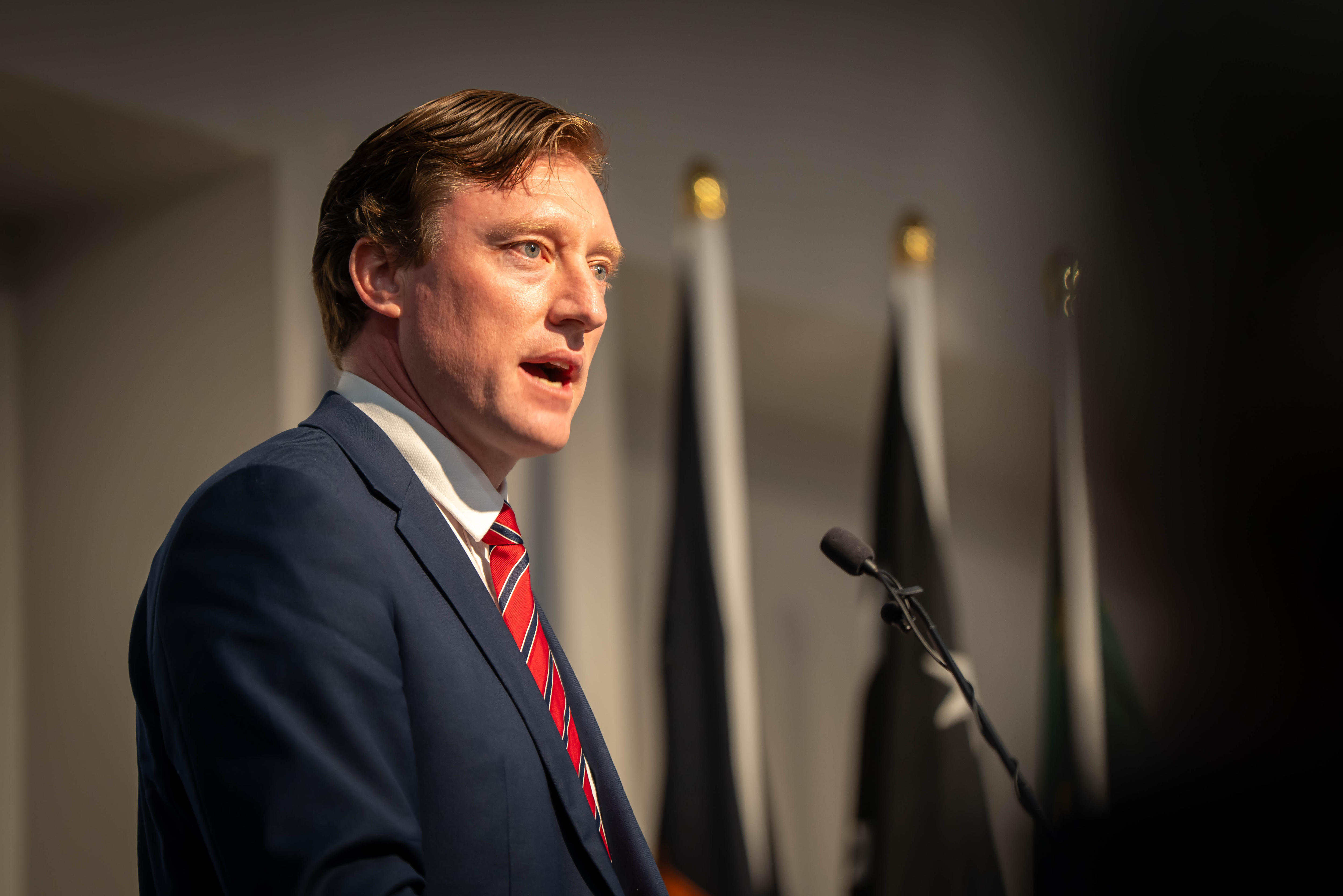 Ed Smelt wears a suit and tie as he speaks into a microphone on a lectern.