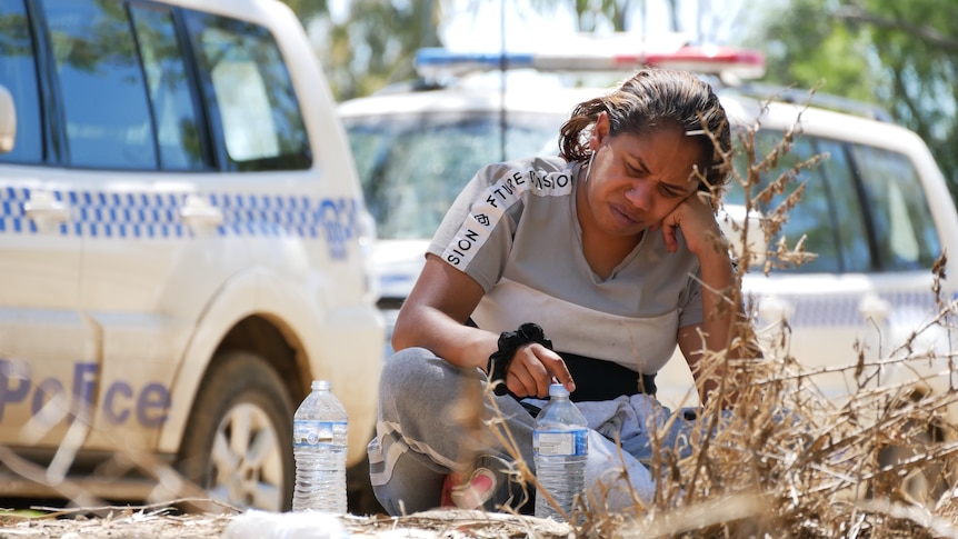 An Indigenous woman sitting cross-legged in front of some police vehicles.